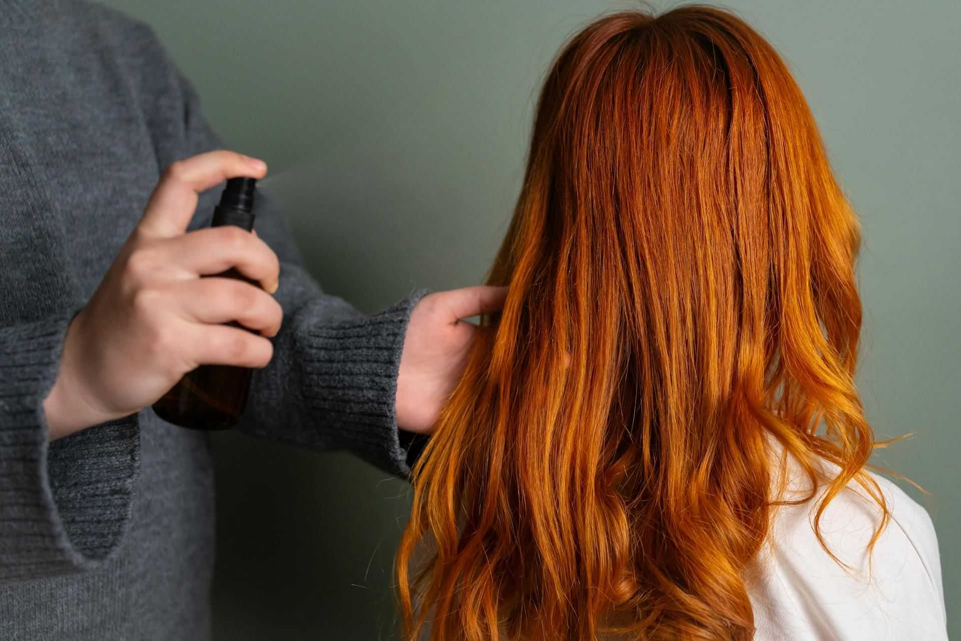 Person spraying product on long, wavy red hair against a green background.